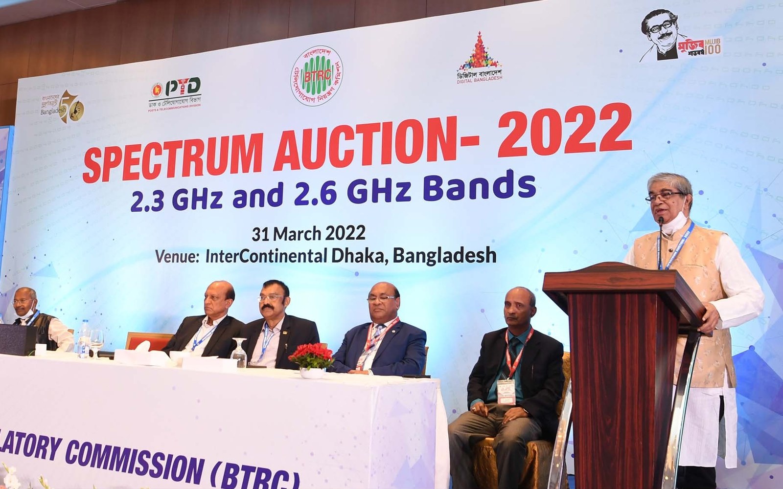 Man speaking at a podium beside seated officials at the Spectrum Auction 2022 event
