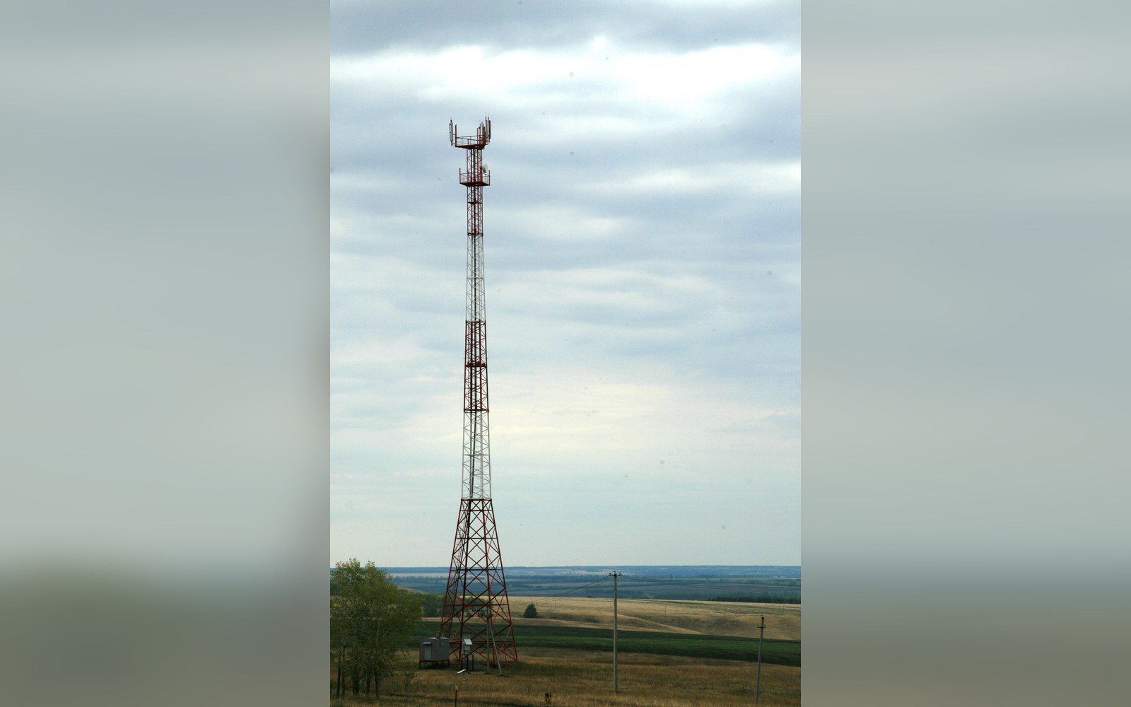 A tall lattice cell tower rises above open farmland under a cloudy sky