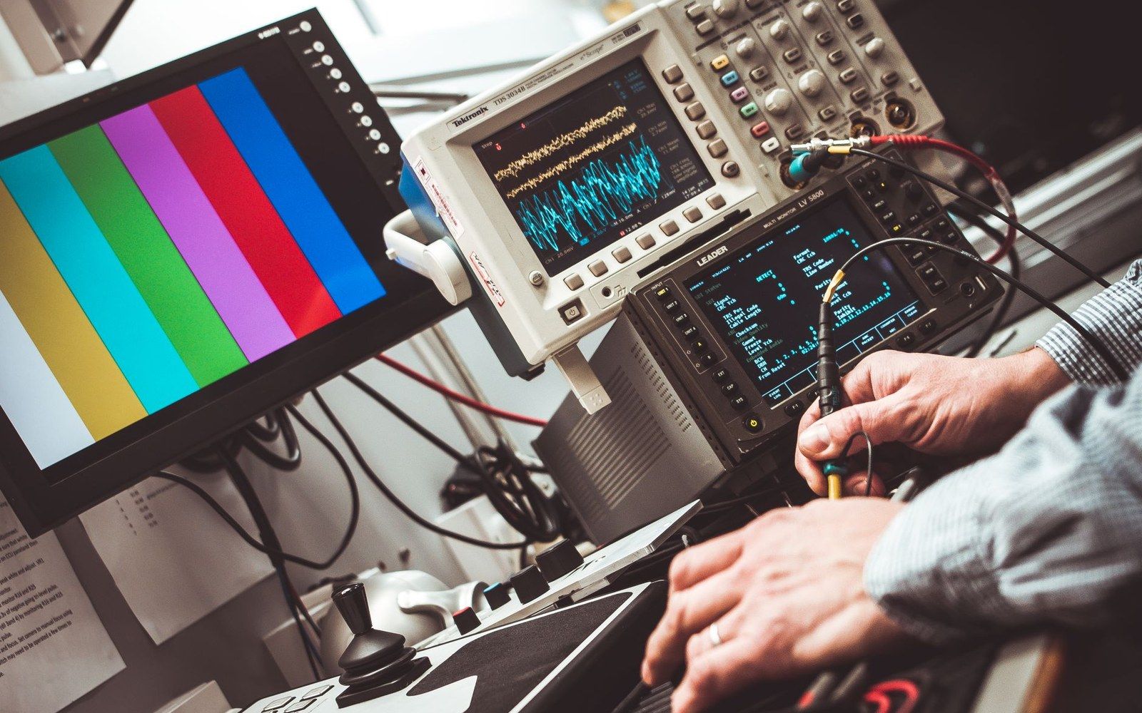 Technician's hands adjust equipment beside a color bar test screen and oscilloscopes